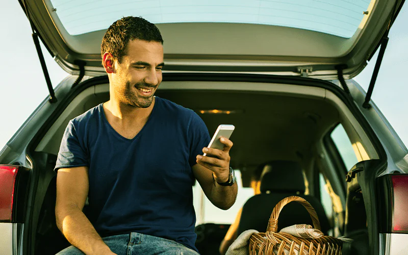 Carro por Assinatura: Homem, com o celular na mão, sentado no porta-malas do carro.